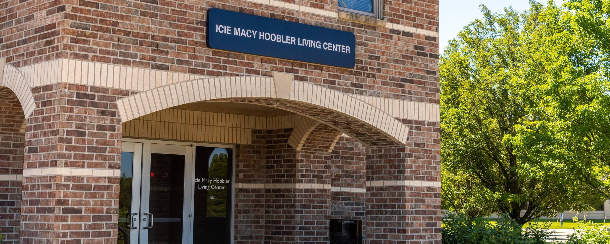 Brick building entrance with a sign reading "Icie Macy Hoobler Living Center" above the doors. A vibrant green tree is on the right, suggesting a peaceful setting.